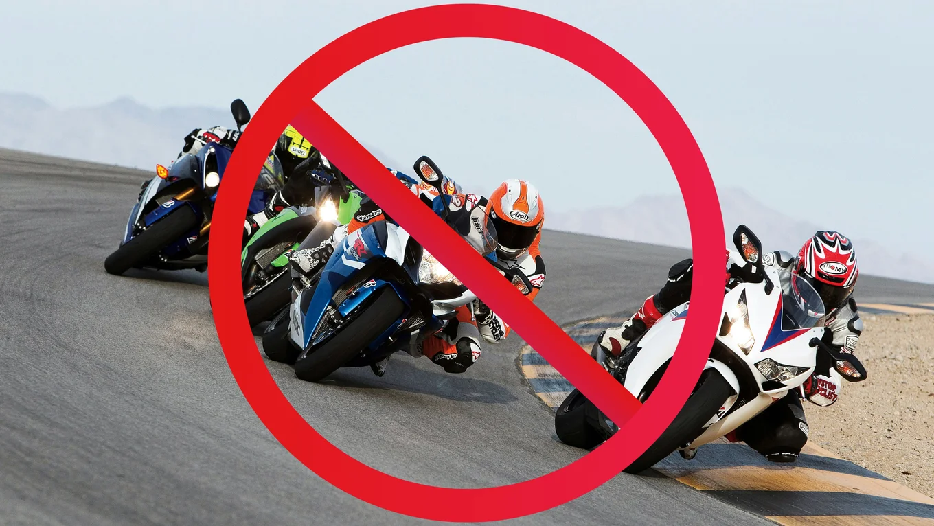 Four motorcycle racers coming around a curve in a desert-like climate, with an outline of mountains in the background. A red stop symbol, a circle with a diagonal line, is imposed on top of the image.